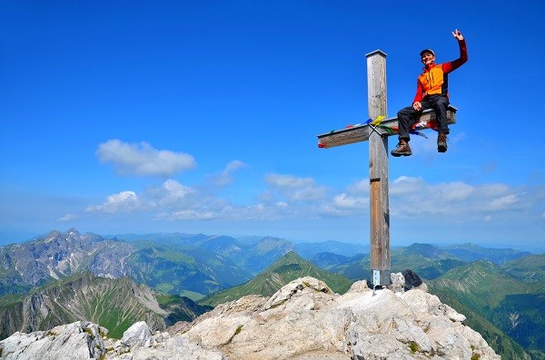 Ein Bergsteiger sitzt auf dem Gipfelkreuz vor blauem Himmel